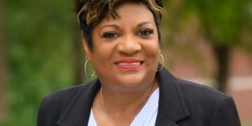 A professional studio portrait of Deb Whitfield, the Mayor of Lawrence, Indiana. She is a Black woman with a warm, confident smile and short, styled dark hair with golden-blonde highlights. She is wearing a black blazer over a white draped-neck blouse, accented by silver hoop earrings and a delicate silver necklace. The background is a soft-focus green landscape with trees, suggesting an outdoor setting with even, natural lighting.