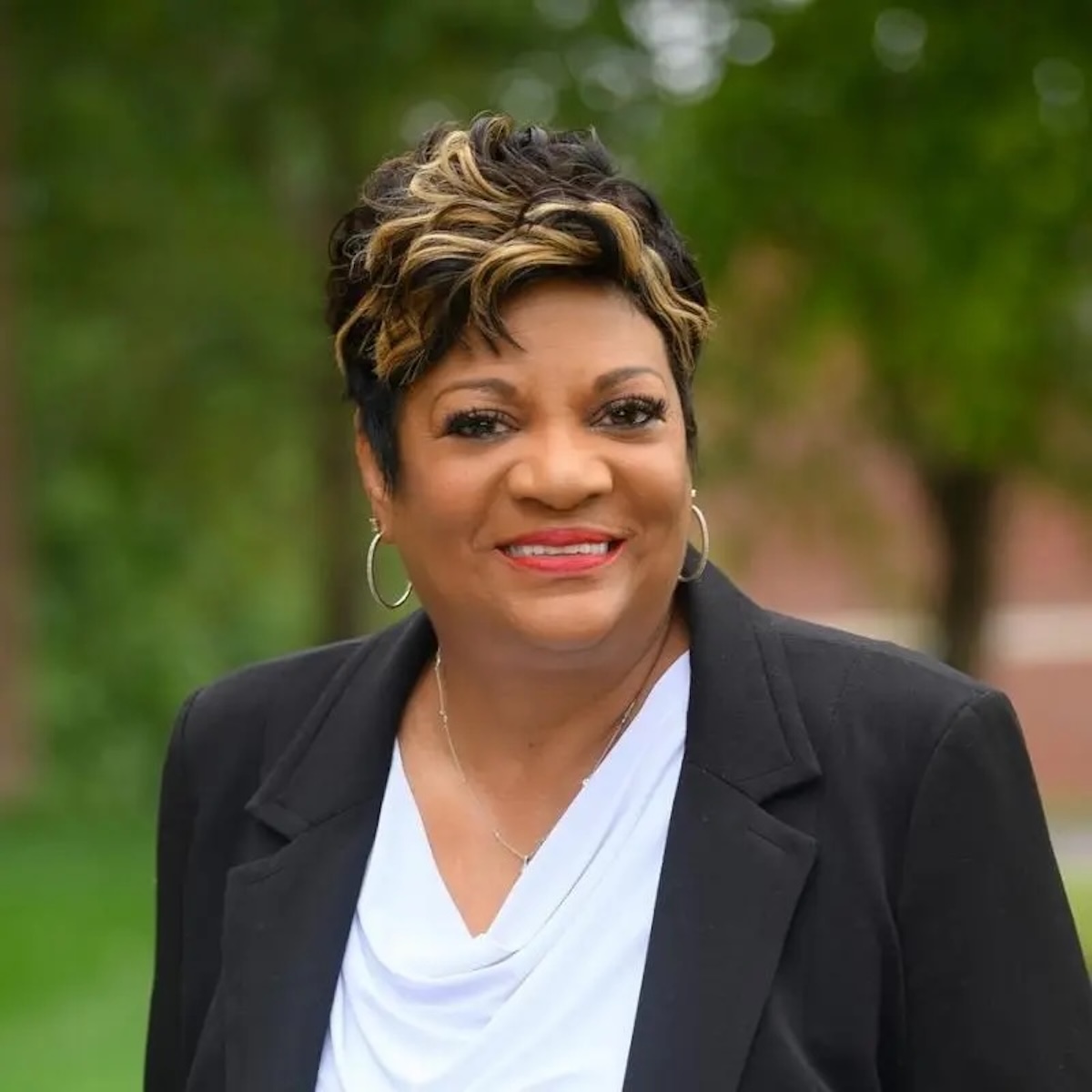 A professional studio portrait of Deb Whitfield, the Mayor of Lawrence, Indiana. She is a Black woman with a warm, confident smile and short, styled dark hair with golden-blonde highlights. She is wearing a black blazer over a white draped-neck blouse, accented by silver hoop earrings and a delicate silver necklace. The background is a soft-focus green landscape with trees, suggesting an outdoor setting with even, natural lighting.