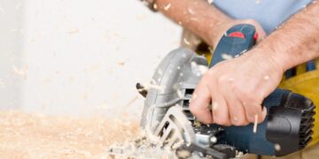 A close-up action shot of a person using a circular saw to cut through a piece of wood. Sawdust and wood chips fly through the air as the blade makes its way across the surface. The person's hands are firmly gripping the tool, and a yellow tool belt is visible at their waist.