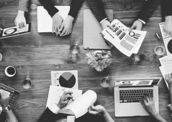 A black-and-white, top-down view of a collaborative business meeting around a wooden conference table. Several professionals are seen working with various tools, including a laptop, a tablet, and printed reports featuring colorful pie charts and bar graphs. Glasses of water and pens are scattered across the table, emphasizing a busy, productive workspace.
