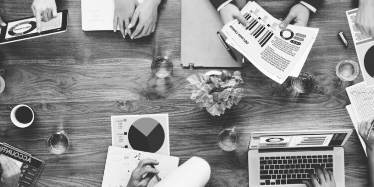 A black-and-white, top-down view of a collaborative business meeting around a wooden conference table. Several professionals are seen working with various tools, including a laptop, a tablet, and printed reports featuring colorful pie charts and bar graphs. Glasses of water and pens are scattered across the table, emphasizing a busy, productive workspace.