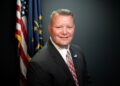 A professional studio headshot of Scott Willis, the Mayor of Westfield, Indiana. He is a man with short brown hair and a warm smile, wearing a dark gray suit, a crisp white button-down shirt, and a patterned red and white tie. On his left lapel, he wears a small pin with the City of Westfield logo. Behind him, the United States flag and the Indiana state flag are displayed on poles against a dark charcoal-colored background.