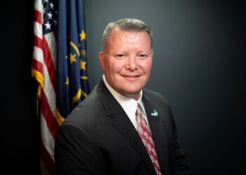 A professional studio headshot of Scott Willis, the Mayor of Westfield, Indiana. He is a man with short brown hair and a warm smile, wearing a dark gray suit, a crisp white button-down shirt, and a patterned red and white tie. On his left lapel, he wears a small pin with the City of Westfield logo. Behind him, the United States flag and the Indiana state flag are displayed on poles against a dark charcoal-colored background.