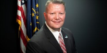 A professional studio headshot of Scott Willis, the Mayor of Westfield, Indiana. He is a man with short brown hair and a warm smile, wearing a dark gray suit, a crisp white button-down shirt, and a patterned red and white tie. On his left lapel, he wears a small pin with the City of Westfield logo. Behind him, the United States flag and the Indiana state flag are displayed on poles against a dark charcoal-colored background.