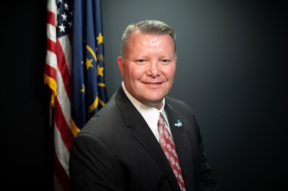 A professional studio headshot of Scott Willis, the Mayor of Westfield, Indiana. He is a man with short brown hair and a warm smile, wearing a dark gray suit, a crisp white button-down shirt, and a patterned red and white tie. On his left lapel, he wears a small pin with the City of Westfield logo. Behind him, the United States flag and the Indiana state flag are displayed on poles against a dark charcoal-colored background.