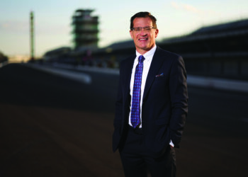 professional portrait of Doug Boles, the President and CEO of Penske Entertainment Corp. and IndyCar. He is a man with short brown hair and glasses, smiling warmly at the camera. He is dressed in a sharp black suit with a patterned blue and purple tie. He is standing on the main straightaway of the Indianapolis Motor Speedway, with his hands in his pockets. The iconic Pagoda and the empty grandstands are visible in the soft-focus background under a warm, late-afternoon sky.