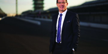 professional portrait of Doug Boles, the President and CEO of Penske Entertainment Corp. and IndyCar. He is a man with short brown hair and glasses, smiling warmly at the camera. He is dressed in a sharp black suit with a patterned blue and purple tie. He is standing on the main straightaway of the Indianapolis Motor Speedway, with his hands in his pockets. The iconic Pagoda and the empty grandstands are visible in the soft-focus background under a warm, late-afternoon sky.