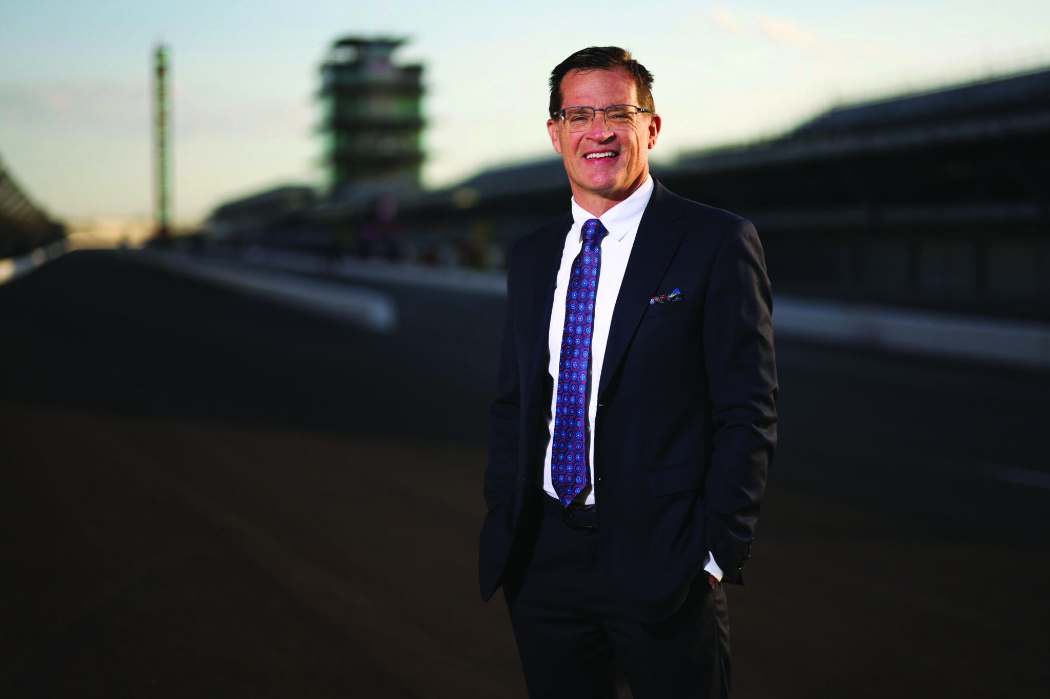 professional portrait of Doug Boles, the President and CEO of Penske Entertainment Corp. and IndyCar. He is a man with short brown hair and glasses, smiling warmly at the camera. He is dressed in a sharp black suit with a patterned blue and purple tie. He is standing on the main straightaway of the Indianapolis Motor Speedway, with his hands in his pockets. The iconic Pagoda and the empty grandstands are visible in the soft-focus background under a warm, late-afternoon sky.