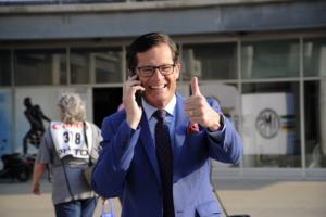 A professional portrait of Doug Boles, the President of the Indianapolis Motor Speedway (IMS) and IndyCar. He is a man with short, dark hair and glasses, smiling broadly while talking on a smartphone and giving a celebratory "thumbs-up" to the camera. He is dressed in a sharp blue windowpane-check suit with a patterned blue and red tie and a red pocket square. He is standing outdoors at the Speedway, with the iconic IMS logo visible on a white pillar in the soft-focus background. To the left, a photographer in a gray "PHOTO" vest is partially visible.