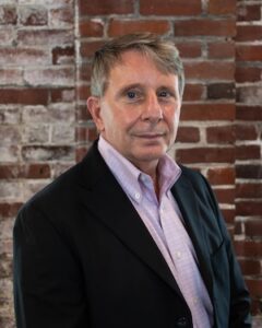 A professional headshot of a smiling man with short, light brown hair. He is wearing a black blazer over a light pink button-down shirt, standing in front of a rustic red and white brick wall.