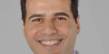 A professional headshot of Dr. Kalid Adab, a hematologist and oncologist. He is smiling warmly, wearing a white lab coat over a light blue collared shirt and a dark patterned tie, set against a neutral, light gray background.