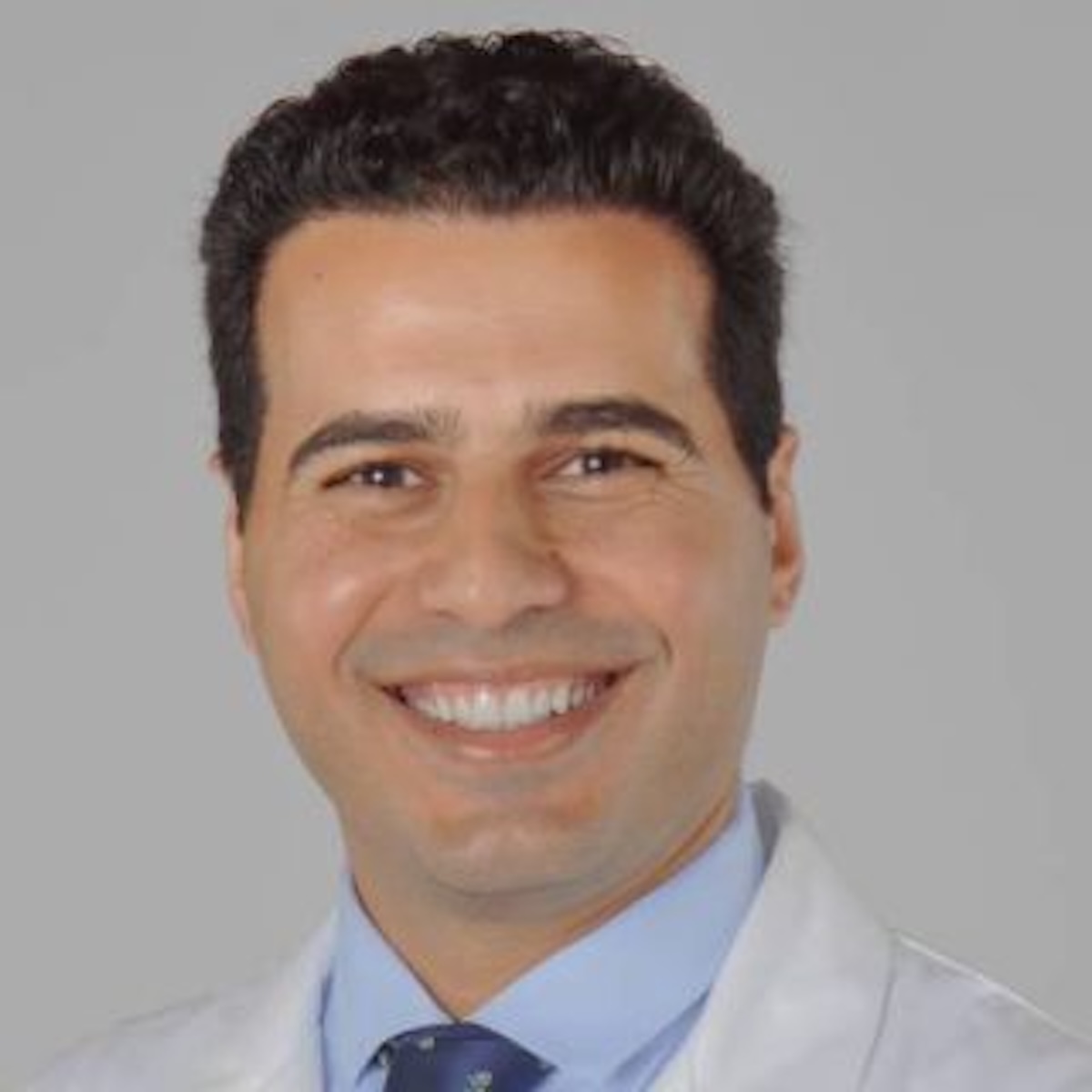 A professional headshot of Dr. Kalid Adab, a hematologist and oncologist. He is smiling warmly, wearing a white lab coat over a light blue collared shirt and a dark patterned tie, set against a neutral, light gray background.