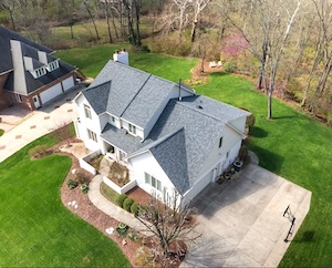 An aerial drone shot showcases a large, two-story white house with a newly installed dark grey shingle roof. The home features several gables, a white chimney, and a multi-car driveway with a basketball hoop. The surrounding property is meticulously landscaped with lush green grass, a curved stone walkway leading to the front entrance, and a dense wooded area in the background under bright, natural daylight.