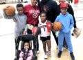 A family of seven poses for a photo on an indoor basketball court, likely at a Center Grove youth league event in Greenwood, Indiana. A man in a black "CG" logo hoodie stands in the back, flanked by two women—one in a red Indiana University sweatshirt and the other in a black top. In front of them, three children are geared up for play: a boy in a gray hoodie holding a Spalding basketball, a girl in a white "Center Grove" jersey (number 7) holding "The Rock" basketball and a red tumbler, and a boy in a light blue hoodie and red cap also holding a basketball. In the foreground, a baby in a pink outfit sits in a black stroller. The background shows a brightly lit gymnasium with other children practicing.