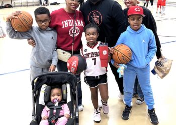 A family of seven poses for a photo on an indoor basketball court, likely at a Center Grove youth league event in Greenwood, Indiana. A man in a black "CG" logo hoodie stands in the back, flanked by two women—one in a red Indiana University sweatshirt and the other in a black top. In front of them, three children are geared up for play: a boy in a gray hoodie holding a Spalding basketball, a girl in a white "Center Grove" jersey (number 7) holding "The Rock" basketball and a red tumbler, and a boy in a light blue hoodie and red cap also holding a basketball. In the foreground, a baby in a pink outfit sits in a black stroller. The background shows a brightly lit gymnasium with other children practicing.