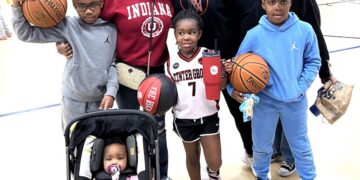 A family of seven poses for a photo on an indoor basketball court, likely at a Center Grove youth league event in Greenwood, Indiana. A man in a black "CG" logo hoodie stands in the back, flanked by two women—one in a red Indiana University sweatshirt and the other in a black top. In front of them, three children are geared up for play: a boy in a gray hoodie holding a Spalding basketball, a girl in a white "Center Grove" jersey (number 7) holding "The Rock" basketball and a red tumbler, and a boy in a light blue hoodie and red cap also holding a basketball. In the foreground, a baby in a pink outfit sits in a black stroller. The background shows a brightly lit gymnasium with other children practicing.
