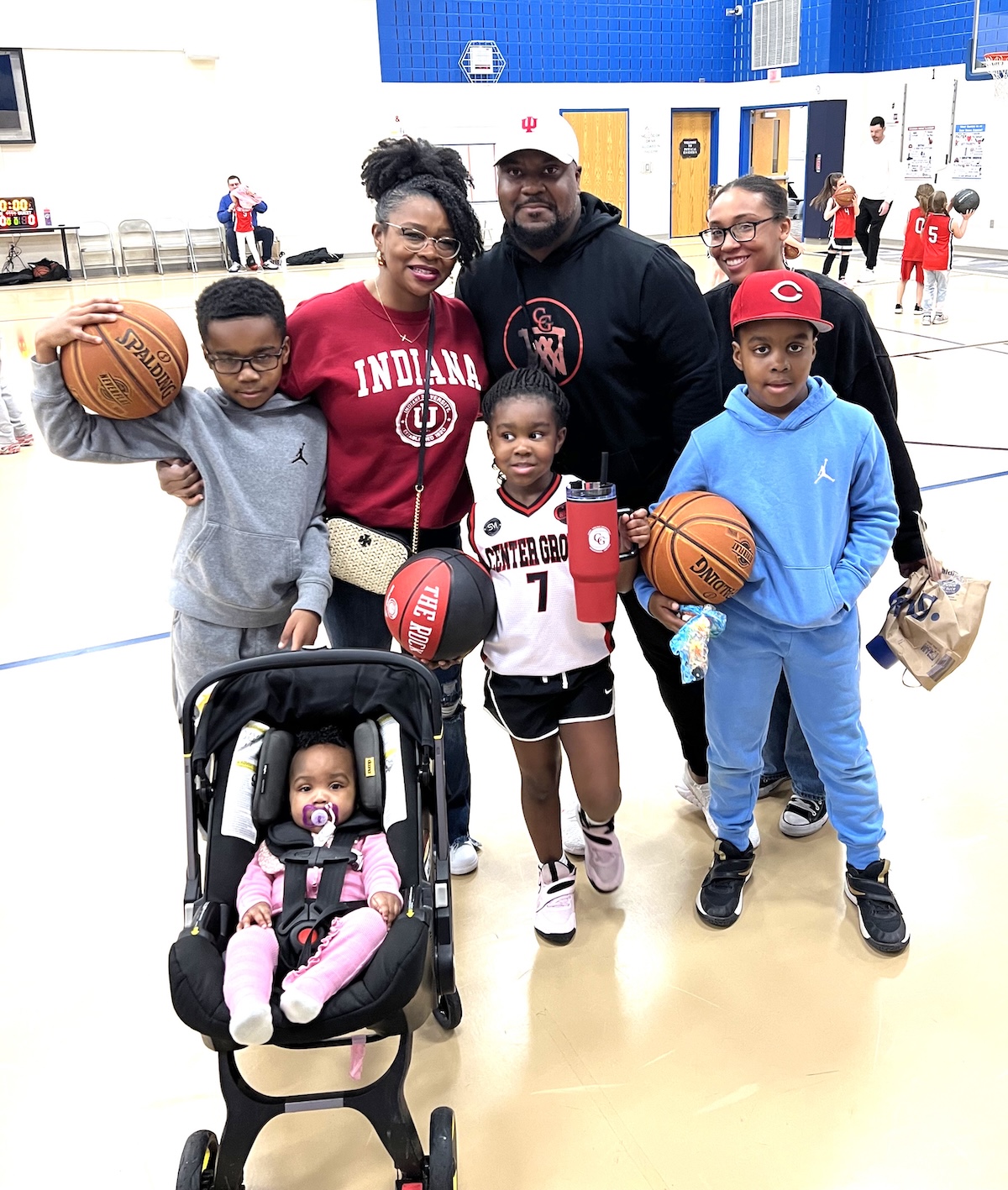 A family of seven poses for a photo on an indoor basketball court, likely at a Center Grove youth league event in Greenwood, Indiana. A man in a black "CG" logo hoodie stands in the back, flanked by two women—one in a red Indiana University sweatshirt and the other in a black top. In front of them, three children are geared up for play: a boy in a gray hoodie holding a Spalding basketball, a girl in a white "Center Grove" jersey (number 7) holding "The Rock" basketball and a red tumbler, and a boy in a light blue hoodie and red cap also holding a basketball. In the foreground, a baby in a pink outfit sits in a black stroller. The background shows a brightly lit gymnasium with other children practicing.