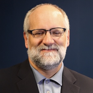 A professional headshot of a smiling man with a white and grey beard and glasses. He is wearing a dark suit jacket over a light blue collared shirt, set against a solid dark blue background.