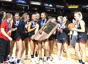 A joyous group of teenage girls, members of the Center Grove Trojans basketball team, celebrate their victory at the 2026 IHSAA Girls Basketball State Finals. They are huddled together on the court at Gainbridge Fieldhouse, wearing state finalist medals around their necks. Two players in the center triumphantly hold a large wooden championship trophy, while a player on the right holds a basketball. They are dressed in black and maroon team uniforms and are laughing and cheering. The background shows the blurred lights and stands of the arena.