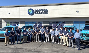 The Raptor Roofing team, a group of fifteen men and women, poses for a group photo in front of their office building. They are dressed in coordinating navy blue and grey company shirts and khakis. The building features a large "RAPTOR ROOFING" sign with a blue and green "RR" logo. Several bright blue Ford F-150 company pickup trucks are parked in a row behind the team under a clear, sunny sky.
