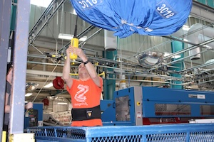 A man in a bright orange t-shirt and a black back-support belt operates a yellow overhead control in an industrial laundry facility. Above him, a large blue bag filled with linens is suspended from a monorail system. In the background, heavy-duty blue laundry machinery and metal overhead rails are visible.
