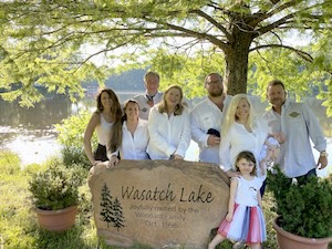 A bright, outdoor group portrait of seven adults and one young girl posing behind a large commemorative stone at Wasatch Lake. The stone features a carved pine tree and the words "Wasatch Lake." Most of the adults are wearing coordinated white or light-colored shirts. They are gathered under the shade of a large, leafy tree with a shimmering lake and lush greenery visible in the background. The young girl in the foreground wears a white top and a colorful, striped skirt.
