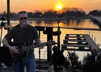 A man with short brown hair and sunglasses, identified as Steve Honeycutt, "The Singing Firefighter," performs a live acoustic set on an outdoor wooden deck at sunset. He is playing a dark-colored acoustic guitar and singing into a microphone. The background features a breathtaking orange and yellow sunset over a calm lake, with a long bridge and distant trees visible on the horizon. The deck is equipped with professional audio equipment, including a spare electric guitar on a stand and a pedalboard on the floor.