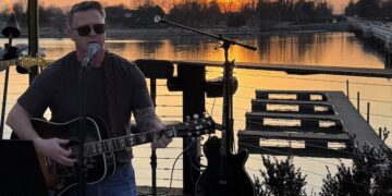 A man with short brown hair and sunglasses, identified as Steve Honeycutt, "The Singing Firefighter," performs a live acoustic set on an outdoor wooden deck at sunset. He is playing a dark-colored acoustic guitar and singing into a microphone. The background features a breathtaking orange and yellow sunset over a calm lake, with a long bridge and distant trees visible on the horizon. The deck is equipped with professional audio equipment, including a spare electric guitar on a stand and a pedalboard on the floor.