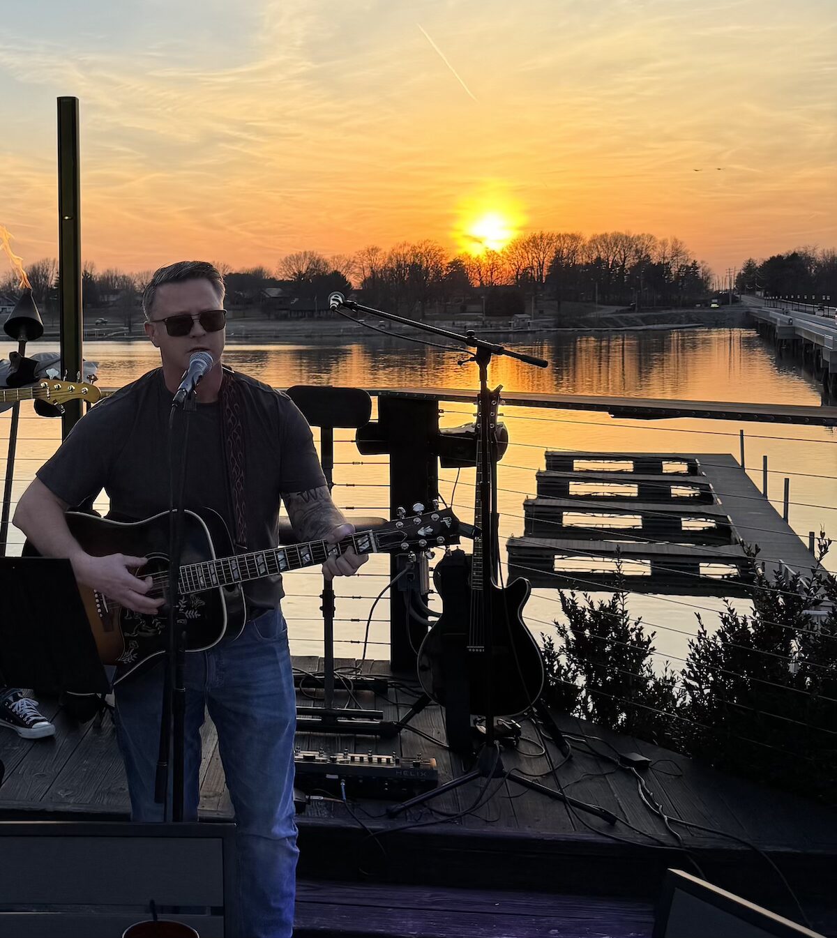 A man with short brown hair and sunglasses, identified as Steve Honeycutt, "The Singing Firefighter," performs a live acoustic set on an outdoor wooden deck at sunset. He is playing a dark-colored acoustic guitar and singing into a microphone. The background features a breathtaking orange and yellow sunset over a calm lake, with a long bridge and distant trees visible on the horizon. The deck is equipped with professional audio equipment, including a spare electric guitar on a stand and a pedalboard on the floor.