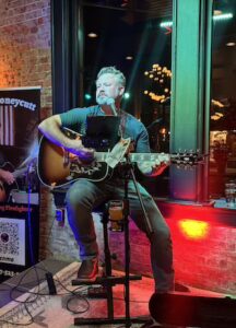 A medium, eye-level shot of Steve Honeycutt, "The Singing Firefighter," performing an acoustic set in a cozy, indoor venue with exposed red brick walls. Steve, a man with graying hair and a beard, is seated on a tall stool, playing a sunburst Gibson acoustic guitar and singing into a microphone. He is wearing a dark teal t-shirt, jeans, and brown boots. To his left, a promotional banner features his name and a QR code for Venmo. The scene is warmly lit by green and red stage lights, with a large window in the background reflecting the warm glow of indoor pendant lights. A pint of beer sits in a holder on his microphone stand.
