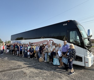 A group of approximately 30 travelers poses for a photo alongside a large, modern Cardinal motorcoach (bus number 1114). The white charter bus features the company's distinctive red and blue "Cardinal" branding and a large stylized logo on its side. The group, likely part of a Dash Getaways tour, is standing on a paved lot under a clear, bright sky.
