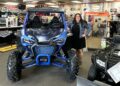 A woman with long dark hair and glasses, wearing a black dress and denim jacket, stands smiling next to a vibrant Matte Navy Blue 2025 Honda Talon 1000X FOX Live Valve side-by-side. The scene is inside the Tom Wood Powersports South dealership in Whiteland, Indiana. The blue off-road vehicle features a prominent black "HONDA" logo, rugged tires, and a protective roll cage. In the background, signs for the "PARTS DEPARTMENT" and "HELMETS" are visible alongside various powersports accessories and another black Honda ATV.