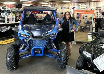 A woman with long dark hair and glasses, wearing a black dress and denim jacket, stands smiling next to a vibrant Matte Navy Blue 2025 Honda Talon 1000X FOX Live Valve side-by-side. The scene is inside the Tom Wood Powersports South dealership in Whiteland, Indiana. The blue off-road vehicle features a prominent black "HONDA" logo, rugged tires, and a protective roll cage. In the background, signs for the "PARTS DEPARTMENT" and "HELMETS" are visible alongside various powersports accessories and another black Honda ATV.