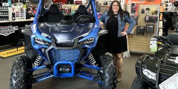 A woman with long dark hair and glasses, wearing a black dress and denim jacket, stands smiling next to a vibrant Matte Navy Blue 2025 Honda Talon 1000X FOX Live Valve side-by-side. The scene is inside the Tom Wood Powersports South dealership in Whiteland, Indiana. The blue off-road vehicle features a prominent black "HONDA" logo, rugged tires, and a protective roll cage. In the background, signs for the "PARTS DEPARTMENT" and "HELMETS" are visible alongside various powersports accessories and another black Honda ATV.
