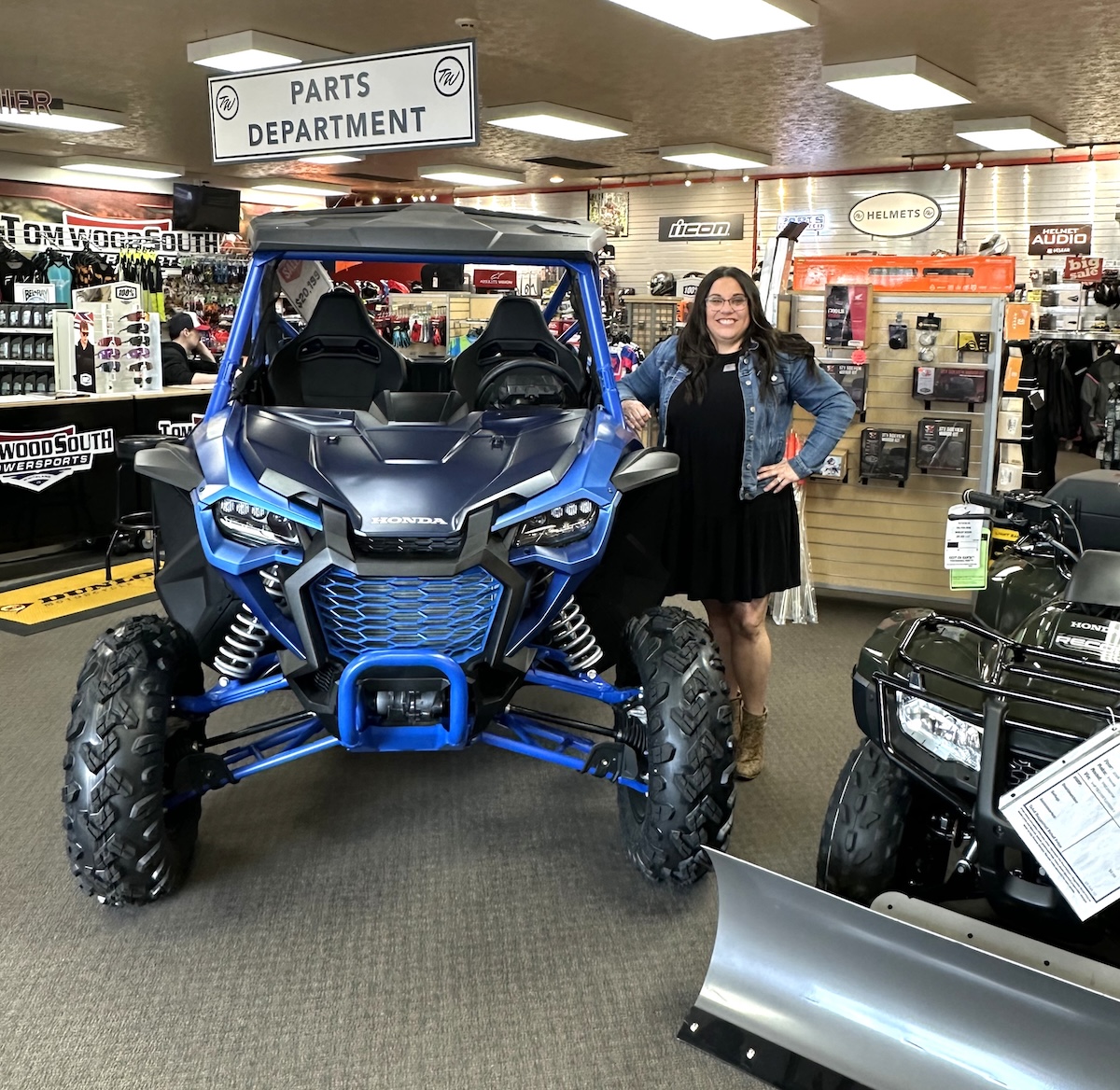 A woman with long dark hair and glasses, wearing a black dress and denim jacket, stands smiling next to a vibrant Matte Navy Blue 2025 Honda Talon 1000X FOX Live Valve side-by-side. The scene is inside the Tom Wood Powersports South dealership in Whiteland, Indiana. The blue off-road vehicle features a prominent black "HONDA" logo, rugged tires, and a protective roll cage. In the background, signs for the "PARTS DEPARTMENT" and "HELMETS" are visible alongside various powersports accessories and another black Honda ATV.