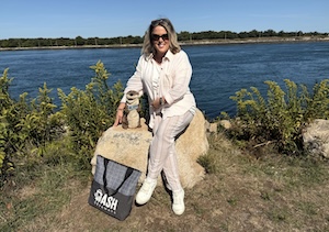 A woman in light-colored clothing and sunglasses sits on a large rock by a wide river, smiling next to a small plush otter. A grey and black Dash Getaways tote bag sits on the ground in front of her. The background features green riverbank foliage and a clear blue sky.