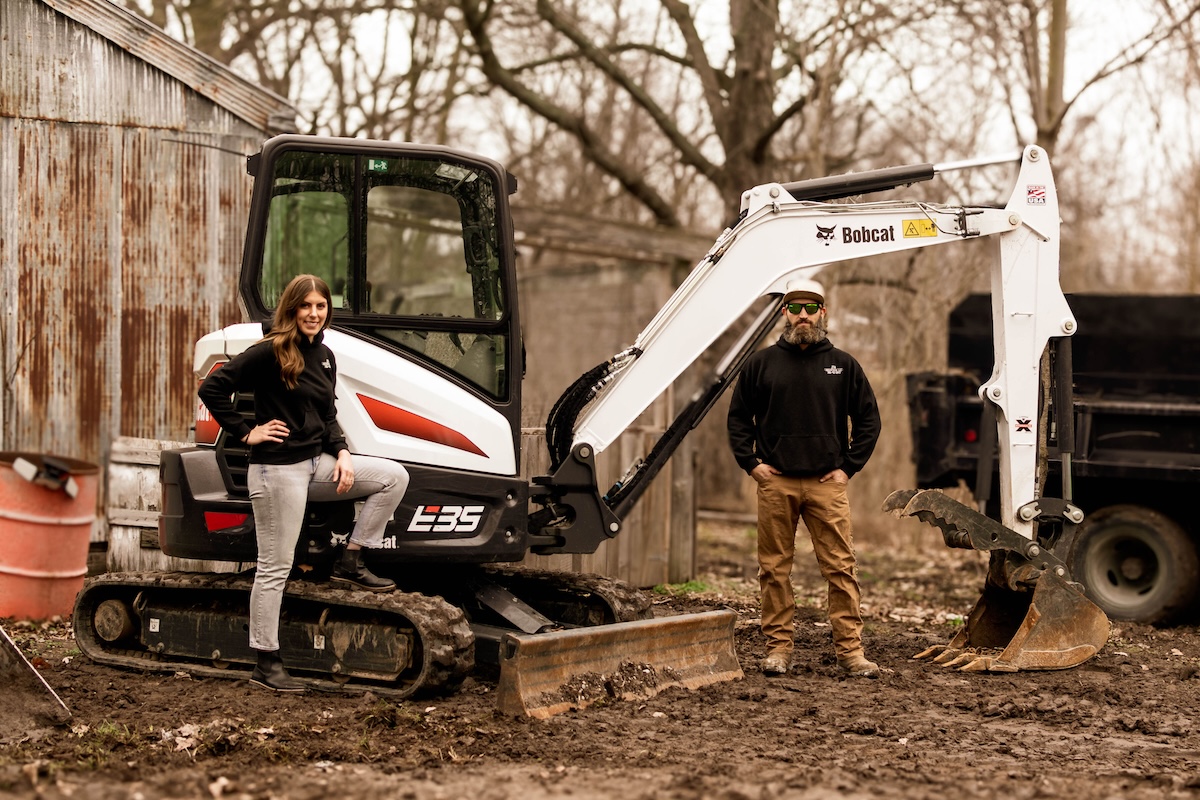 A man and a woman pose confidently with a white and black Bobcat E35 compact excavator in a muddy, wooded area.
