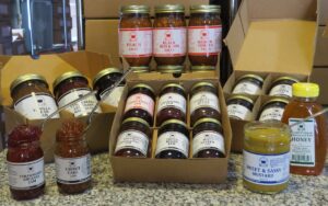 A large collection of artisanal jarred goods, including various jams, salsas, and honey, are displayed on a granite countertop. In the foreground, two open jars of Strawberry Rhubarb Jam and Carrot Cake Jam have spoons resting in them, showcasing their thick textures. Next to them is a jar of Sweet & Sassy Mustard and a large plastic bottle of Raw Honey. The background features several cardboard gift boxes filled with assorted jars such as Peach Salsa, Black Bean & Corn Salsa, Triple Berry Jam, and Hot Pepper Jam. All products feature uniform white labels with red or black text and a circular blue logo.