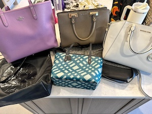 A collection of six designer handbags in various colors and styles is arranged on a light-colored countertop. The group includes a purple Coach tote, a tan leather satchel with gold hardware, and a cream-colored Michael Kors handbag. Below them are a black Tory Burch tote with a large circular logo, a smaller teal and white geometric pattern bag, and a black crossbody purse. A white travel tumbler is partially visible in the background.