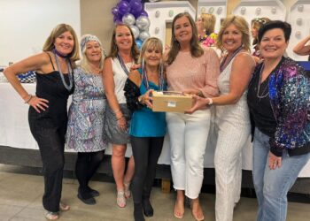 Seven women smile together at an event, with two in the center holding a small cardboard bakery box. They are dressed in a mix of festive, sparkly, and patterned outfits, including a black sequined jumpsuit, a retro geometric-print dress, and a blue sequined bomber jacket. Many of the women are wearing purple and silver bead necklaces. In the background, purple and silver balloons and large marquee-style letters with lights are visible.