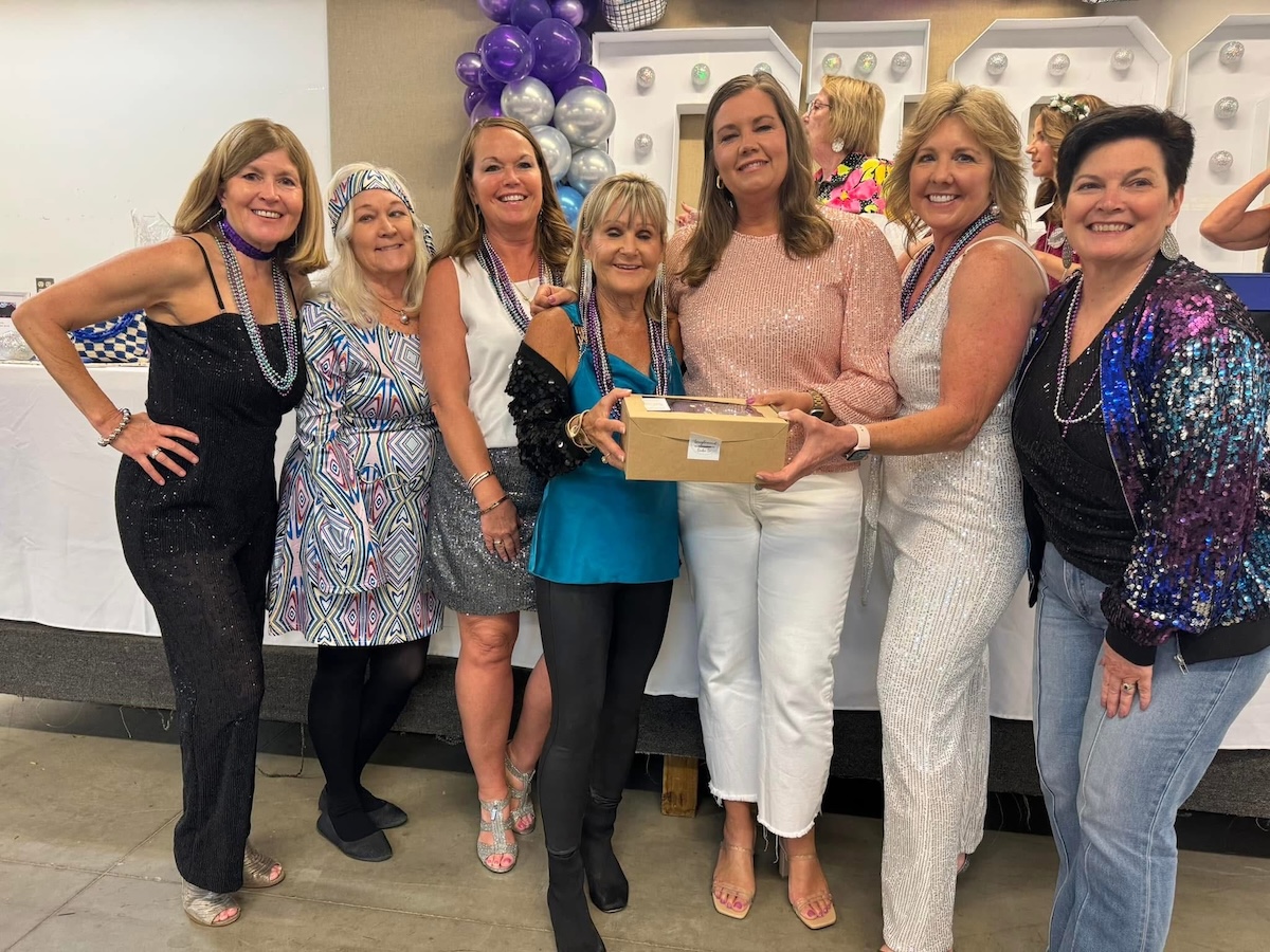 Seven women smile together at an event, with two in the center holding a small cardboard bakery box. They are dressed in a mix of festive, sparkly, and patterned outfits, including a black sequined jumpsuit, a retro geometric-print dress, and a blue sequined bomber jacket. Many of the women are wearing purple and silver bead necklaces. In the background, purple and silver balloons and large marquee-style letters with lights are visible.