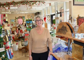 A smiling woman with grey hair and glasses stands in the center of a brightly lit gift shop. She wears a tan v-neck sweater and black pants. The shop is filled with various items, including Kentucky-themed decor, a decorated Christmas tree, colorful socks, and shelves stocked with glassware and apparel. To her right, a wooden display features "Kentucky Bourbon" branding.