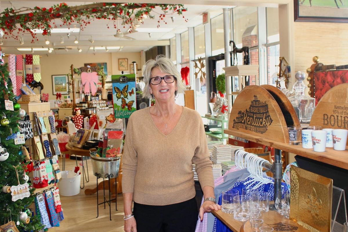 A smiling woman with grey hair and glasses stands in the center of a brightly lit gift shop. She wears a tan v-neck sweater and black pants. The shop is filled with various items, including Kentucky-themed decor, a decorated Christmas tree, colorful socks, and shelves stocked with glassware and apparel. To her right, a wooden display features "Kentucky Bourbon" branding.