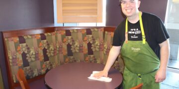 In a bright Panera Bread dining area, a smiling male employee in a green apron, black "It's a New Era at Panera" T-shirt, and a grey cap is cleaning a round purple table with a white cloth. He is standing next to a curved booth with floral-patterned upholstery in shades of purple, orange, and tan.