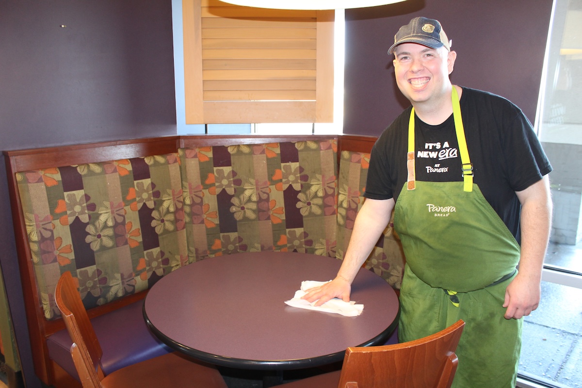In a bright Panera Bread dining area, a smiling male employee in a green apron, black "It's a New Era at Panera" T-shirt, and a grey cap is cleaning a round purple table with a white cloth. He is standing next to a curved booth with floral-patterned upholstery in shades of purple, orange, and tan.