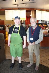 Inside a brightly lit Panera Bread, a young male employee and an older man stand side-by-side, smiling for a photo.
On the left: The employee wears a green Panera apron over a black T-shirt that says "It's a New Era at Panera." He is also wearing a blue baseball cap and black sneakers.
On the right: An older man with a white beard wears a checkered button-down shirt under a navy blue sweater vest, tan trousers, and a red lanyard around his neck.
The background shows the restaurant's service counter, glowing menu boards, and a glimpse of the kitchen area.