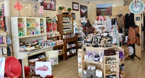 The interior of a brightly lit boutique gift shop featuring light wood floors and cream-colored walls adorned with framed artwork. A white hutch displays colorful bottled lotions and soaps, while a central wooden shelving unit showcases a variety of local artisan products, including jewelry, candles, and skincare. In the background, clothing racks and Kentucky-themed decor are neatly organized throughout the space.