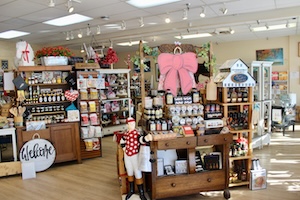 The interior of a brightly lit gift shop filled with various displays of gourmet food items and home decor. In the foreground, a wooden buffet table showcases jars of jams and honey, topped with a large, decorative pink bow. Nearby, a jockey statue in a red-and-white polka dot silks stands next to shelves of Kentucky-themed products. A large round "Welcome" sign sits on the light wood floor amidst the well-organized aisles.