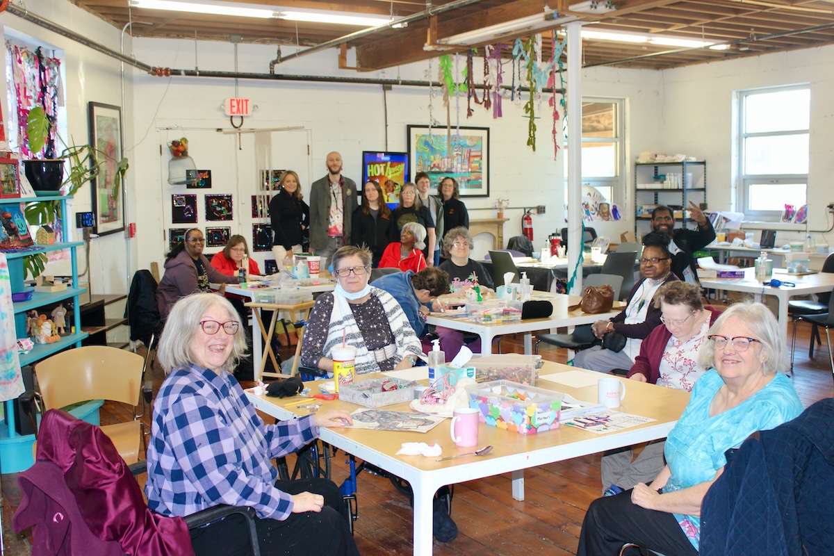 A large, diverse group of people are gathered in a bright, industrial-style art studio with exposed ceiling beams and hardwood floors.