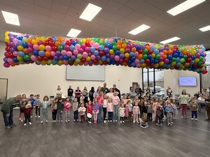 In a large, open room with grey flooring, a diverse group of approximately 30 children and several adults stand together for a group photo. Hanging directly above them is a massive, long net filled with hundreds of vibrant, multi-colored balloons in shades of red, blue, yellow, green, and pink, ready for a balloon drop. The room is brightly lit by rectangular ceiling lights, and large windows are visible in the background.